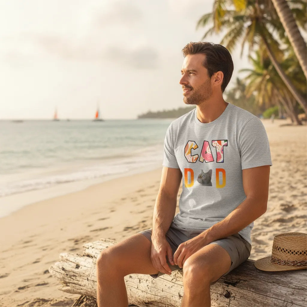 Man sitting on driftwood on a beach wearing the grey CAT DAD t-shirt