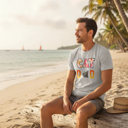 Man sitting on driftwood on a beach wearing the grey CAT DAD t-shirt