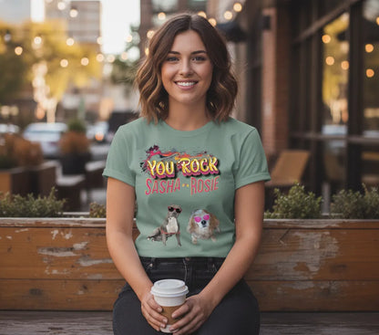 Woman outside a cafe holding a coffee cup, wearing the sage green You Rock t-shirt printed with Sasha and Rosie in sunglasses